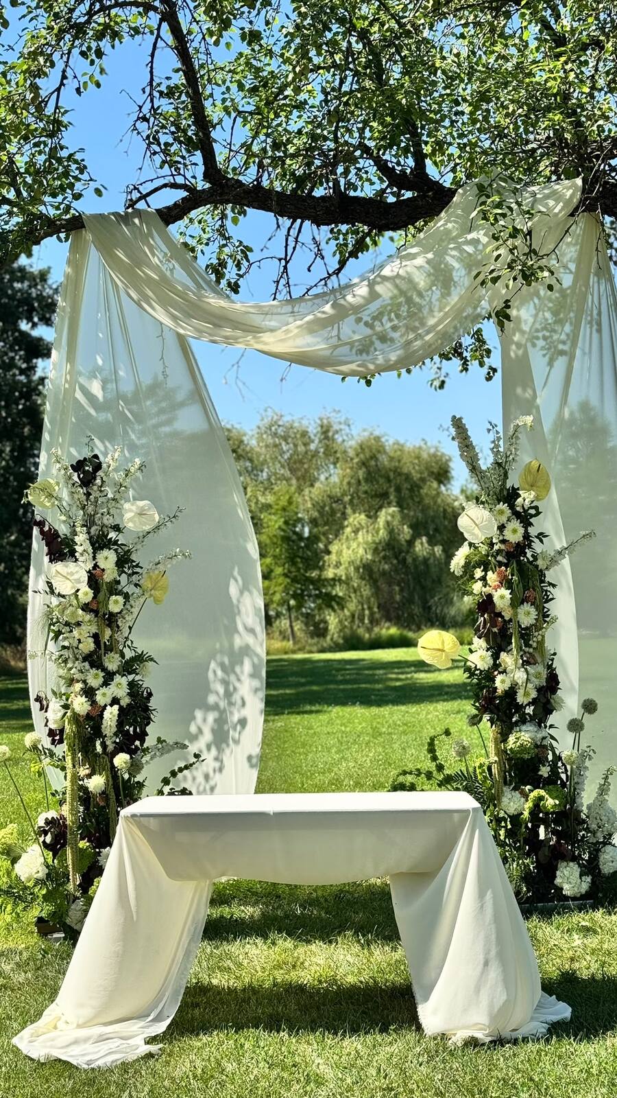 A ceremony arch dressed in ivory drapes and seasonal flowers, prepared under the trees